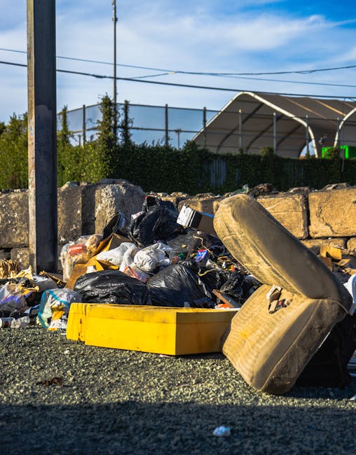 An outdoor scene showing a pile of discarded waste and debris on a paved surface next to a brick wall. The waste includes a wooden slatted bed frame leaning against a wall, a damaged and partially dis