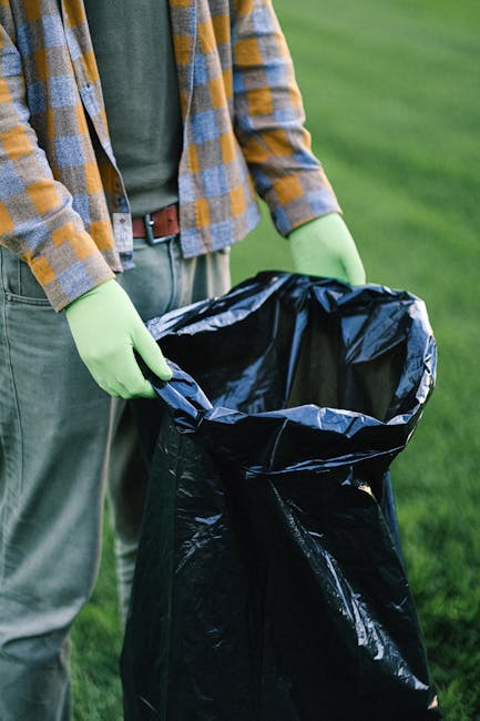 A collection of overflowing rubbish and waste materials is piled on a paved urban sidewalk, near a parking area. The assortment includes numerous black garbage bags, brown cardboard boxes, and crumple