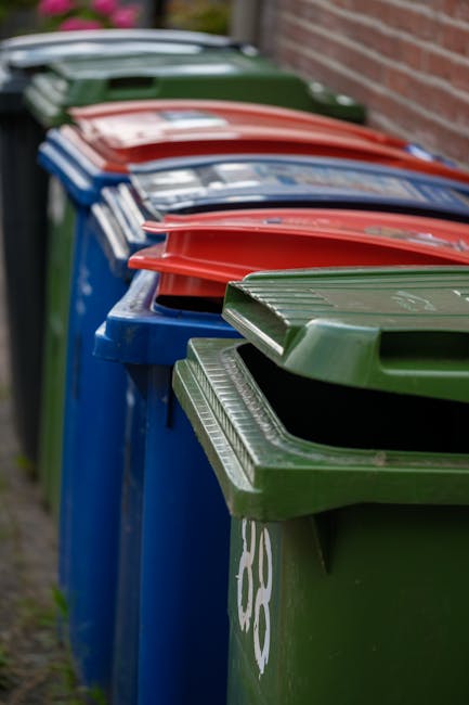 A row of large, rectangular wheelie bins lined up outdoors along a pavement next to a brick wall. The bins are made of durable plastic with textured surfaces on their lids, which have ridged edges for