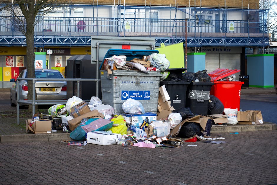 A row of large, rectangular wheelie bins lined up outdoors along a pavement next to a brick wall. The bins are made of durable plastic with textured surfaces on their lids, which have ridged edges for